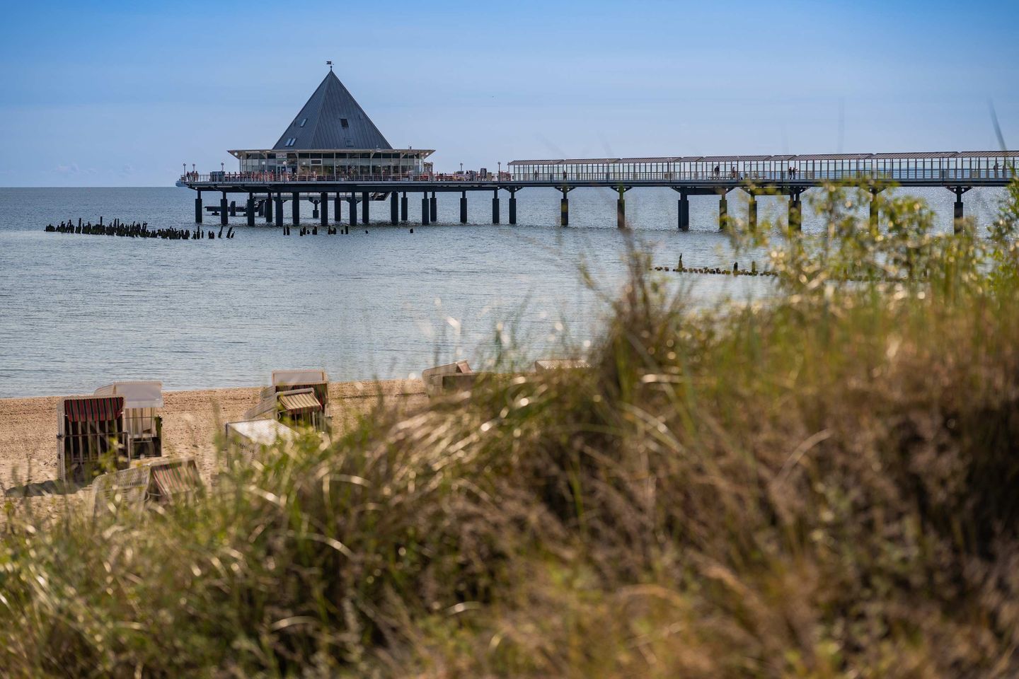 A pier extending into the water with a building at the end, surrounded by calm water and some vegetation in the foreground.