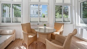 Aurinkoinen huone, jossa on pyöreä lasipöytä, jonka ympärillä on punottuja tuoleja, suuret ikkunat, joissa on sälekaihtimet, ja näkymä ulkona oleviin rakennuksiin.