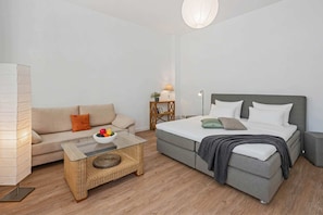 A modern bedroom with a grey bed, a beige sofa, a glass coffee table with fruit, and a wooden side table.