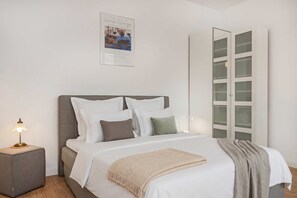 Una cama bien hecha con ropa de cama blanca y gris, una lámpara de noche y un cartel en la pared.