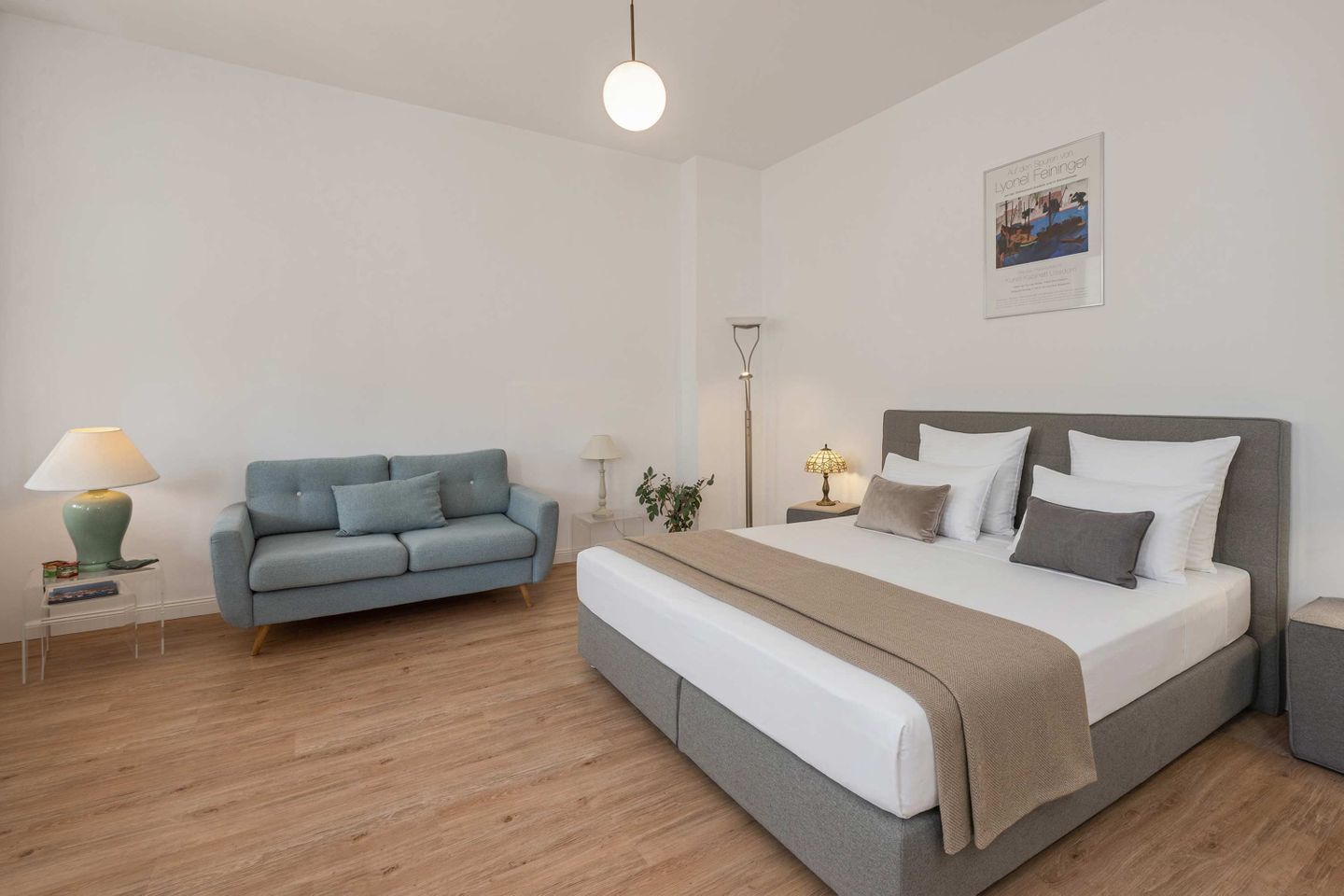 A bedroom with a bed, a grey sofa, a bedside table with a lamp, and a wall-mounted picture.