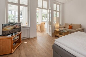 A modern hotel room with a large bed, a TV on a wooden stand, a glass coffee table, a sofa, and a dining area with wicker furniture.