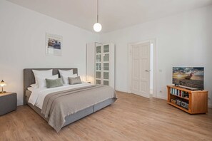 Quarto com cama, mesa de cabeceira, luminária, guarda-roupa e uma televisão sobre um suporte de madeira.