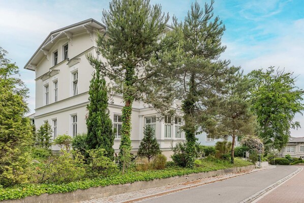 A two-story building with a white facade, surrounded by greenery and trees.