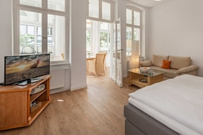 A modern hotel room with a large bed, a TV on a wooden stand, a glass coffee table, a sofa, and a dining area with wicker furniture.