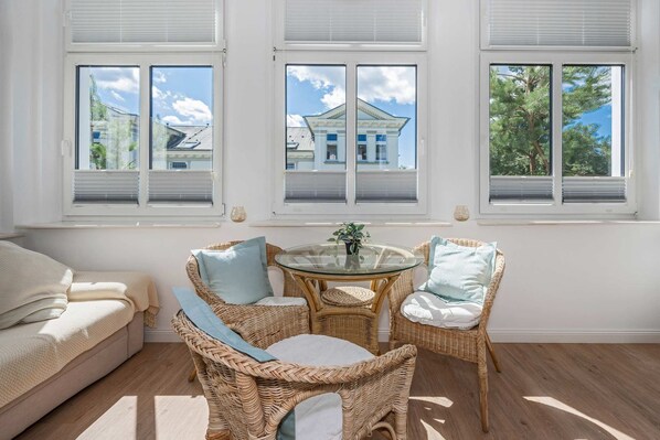 A room with a sofa, wicker chairs, a glass table, and a view of a building through large windows.