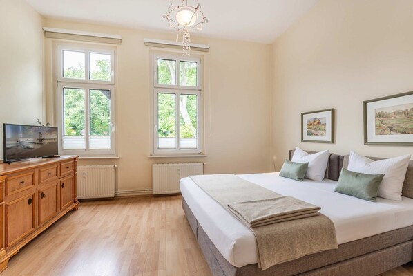 A bedroom with a bed, a TV, a dresser, and two windows with a view of greenery.