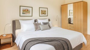 A neatly made bed with white linens and a grey throw blanket, a wooden wardrobe, and two framed landscape paintings on the wall.