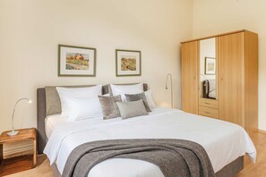 Una cama bien tendida con sábanas blancas y un plaid gris, un armario de madera y dos cuadros con paisajes en la pared.