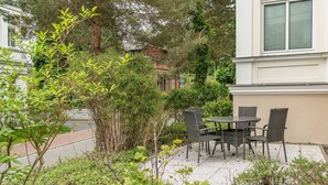 Une terrasse extérieure avec une table et des chaises, entourée de verdure et avec un bâtiment en arrière-plan.
