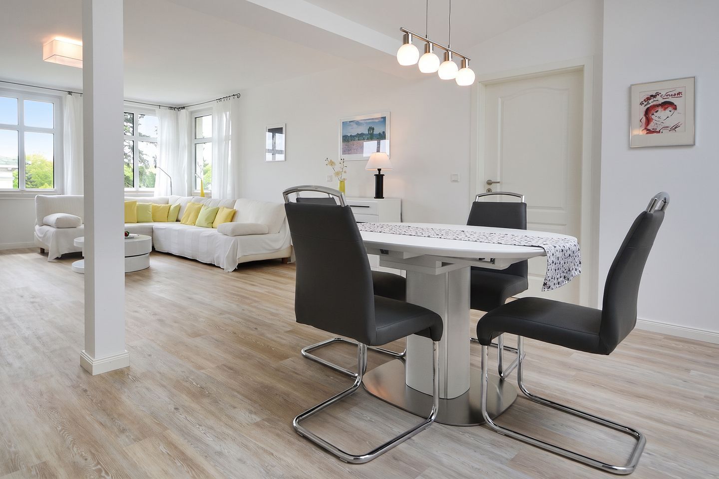 Un espace repas moderne avec une table blanche et des chaises noires, un luminaire suspendu et un canapé blanc en arrière-plan.