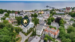 Une vue aérienne d’une ville côtière, avec un mélange de bâtiments résidentiels, une jetée s’avançant dans la mer et un hôtel imposant doté d’un toit rouge.