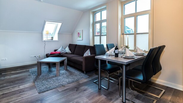 A modern living room with a sofa, coffee table, dining table, and chairs. There are large windows providing natural light. - Altstadt Borkum Apartment 4 Bant - Altstadt Villa Borkum (Borkum)