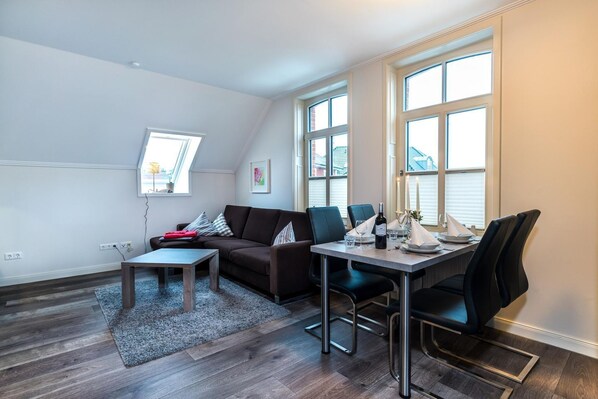 Un salon moderne comprenant un canapé, une table basse, une table à manger et des chaises. De grandes fenêtres laissent entrer la lumière naturelle.