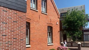 A red brick building with a flat roof and a balcony. There are windows with white frames and a small clock on the wall. - Altstadt Borkum Apartment 4 Bant - Altstadt Villa Borkum (Borkum)
