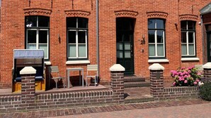 A red brick building with a pitched roof, multiple windows, and a small porch area with outdoor seating. - Altstadt Borkum Apartment 4 Bant - Altstadt Villa Borkum (Borkum)