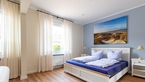 Et soveværelse med en seng, et natbord, en lampe, et vindue med gardiner og et billede på væggen.