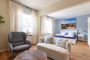 A bedroom with a bed, a grey armchair, a beige sofa, a round table, and a painting on the wall.