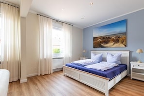A bedroom with a bed, a nightstand, a lamp, a window with curtains, and a painting on the wall.