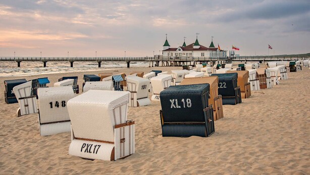 Beachside wicker chairs with numbers, a pier, and a building with green roofs in the background.