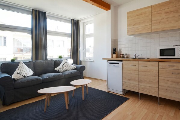 A modern living room with a grey sofa, two round tables, a black rug, a kitchen with wooden cabinets, a white microwave, and a dishwasher.