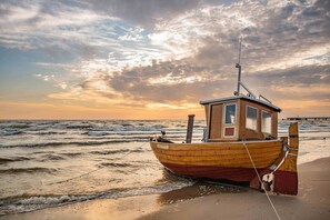 Un bote de madera con cabina, amarrado en una playa arenosa con olas, bajo un cielo parcialmente nublado.
