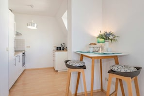 Une cuisine compacte avec des meubles blancs, un plan de travail en bois et un réfrigérateur blanc. On y trouve un coin repas avec une table et deux tabourets, ainsi qu’une petite plante posée sur la table.