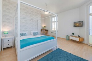 A bedroom with a four-poster bed, a bedside table with a lamp, a small wooden side table, a blue rug, and a TV on the wall. - Apartment Karlskrona in Ahlbeck with balcony, close to the beach - Haus Skanska (Heringsdorf)