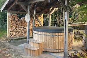 A wooden hot tub with steps, a covered area, and stacked firewood.