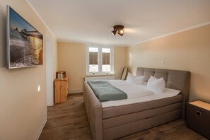 Un dormitorio con cama, ventana con persianas, mesita de noche de madera y un cuadro en la pared.