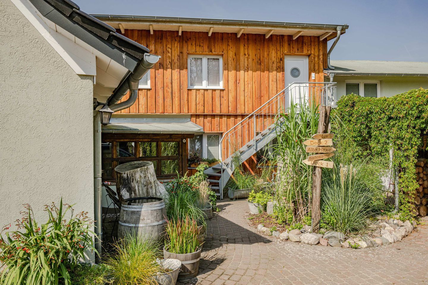 Une maison en bois avec un escalier menant à une terrasse, entourée de plantes en pot et d’un mur de pierres.
