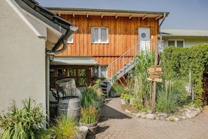Une maison en bois avec un escalier menant à une terrasse, entourée de plantes en pot et d’un mur de pierres.
