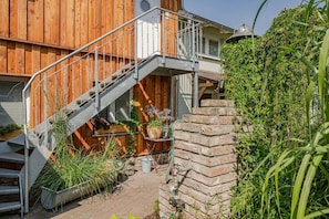 A wooden house with a metal staircase leading to a balcony, surrounded by greenery and a brick wall.