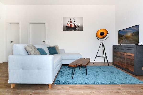 Una sala de estar con un sofá de color claro, un tapete azul, una mesa de centro de madera, una lámpara de pie, un mueble con televisor y un cuadro enmarcado en la pared.