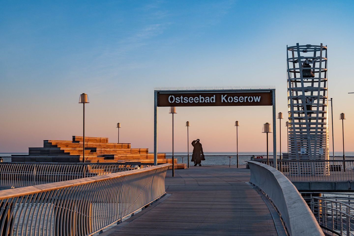 Un muelle con un cartel que dice 'Ostseebad Koserow', bancos, barandillas y una estatua.