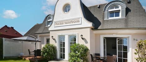 Una casa de dos pisos con un letrero que dice 'Haus Usedore'. Cuenta con un patio con mobiliario exterior, una zona de estar cubierta y un césped bien cuidado.
