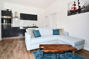 A modern living room with a light-colored sectional sofa, a wooden coffee table, and a kitchen area with dark cabinets and a stove. - Cozy vacation apartment in Koserow with terrace, only 350 m to the beach - House (Koserow)