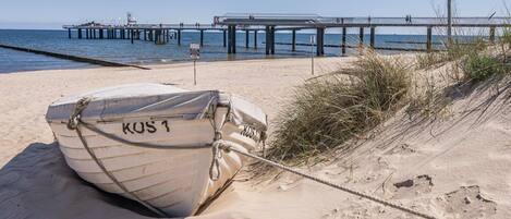 Ein Boot mit dem Namen 'KOS 1' liegt an einem Sandstrand, im Hintergrund ist ein Steg zu sehen.