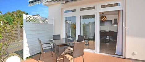 A covered outdoor patio with a table and chairs, a white fence, and a view into the house.