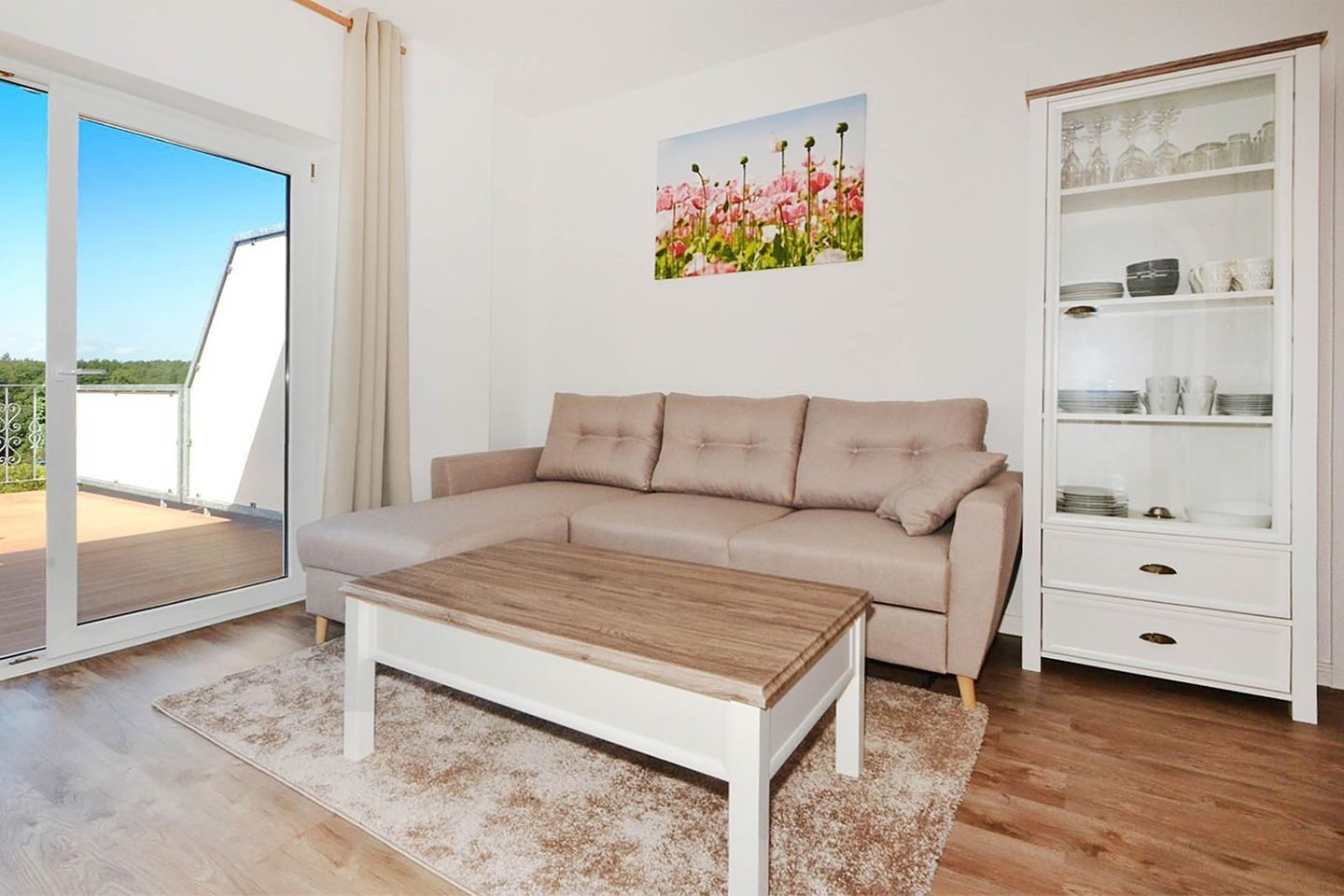 Un salon moderne avec un canapé d’angle beige, une table basse en bois, un meuble vitré contenant de la vaisselle et un tableau représentant des fleurs accroché au mur.