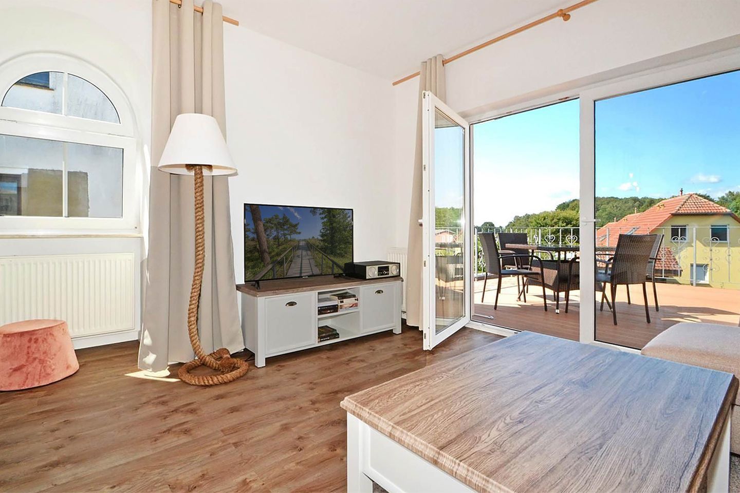 A room with a television, a white cabinet, a wooden floor, a pink ottoman, and a sliding glass door leading to an outdoor area with a table and chairs.