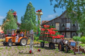 A rural scene with a tractor pulling a trailer of crates, a vintage car, and a wooden building.