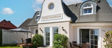 A two-story house with a sign reading 'Haus Usedore.' It features a patio with outdoor furniture, a covered seating area, and a well-maintained lawn.