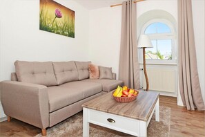 A living room with a beige sofa, a wooden coffee table with a fruit basket, a lamp, and a window with curtains.