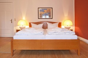 A double bed with white linens, two bedside lamps, a framed artwork above the bed, and a wooden floor.
