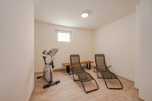Ein Zimmer mit Deckenventilator, einem Heimtrainer, einem Holztisch und zwei Liegesesseln.