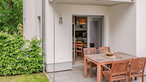 A patio with a wooden table and chairs, a small garden, and a building entrance. - Modern vacation apartment in Heringsdorf with terrace, only 1000 m from the beach (Heringsdorf)