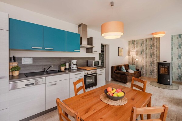 A modern kitchen with teal cabinets, a dining table with chairs, a fruit bowl, and a wood-burning stove.
