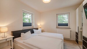 A bedroom with a bed, two bedside tables, a nightstand, a wall-mounted TV, and two windows with blinds. - Modern vacation apartment in Heringsdorf with terrace, only 1000 m from the beach (Heringsdorf)