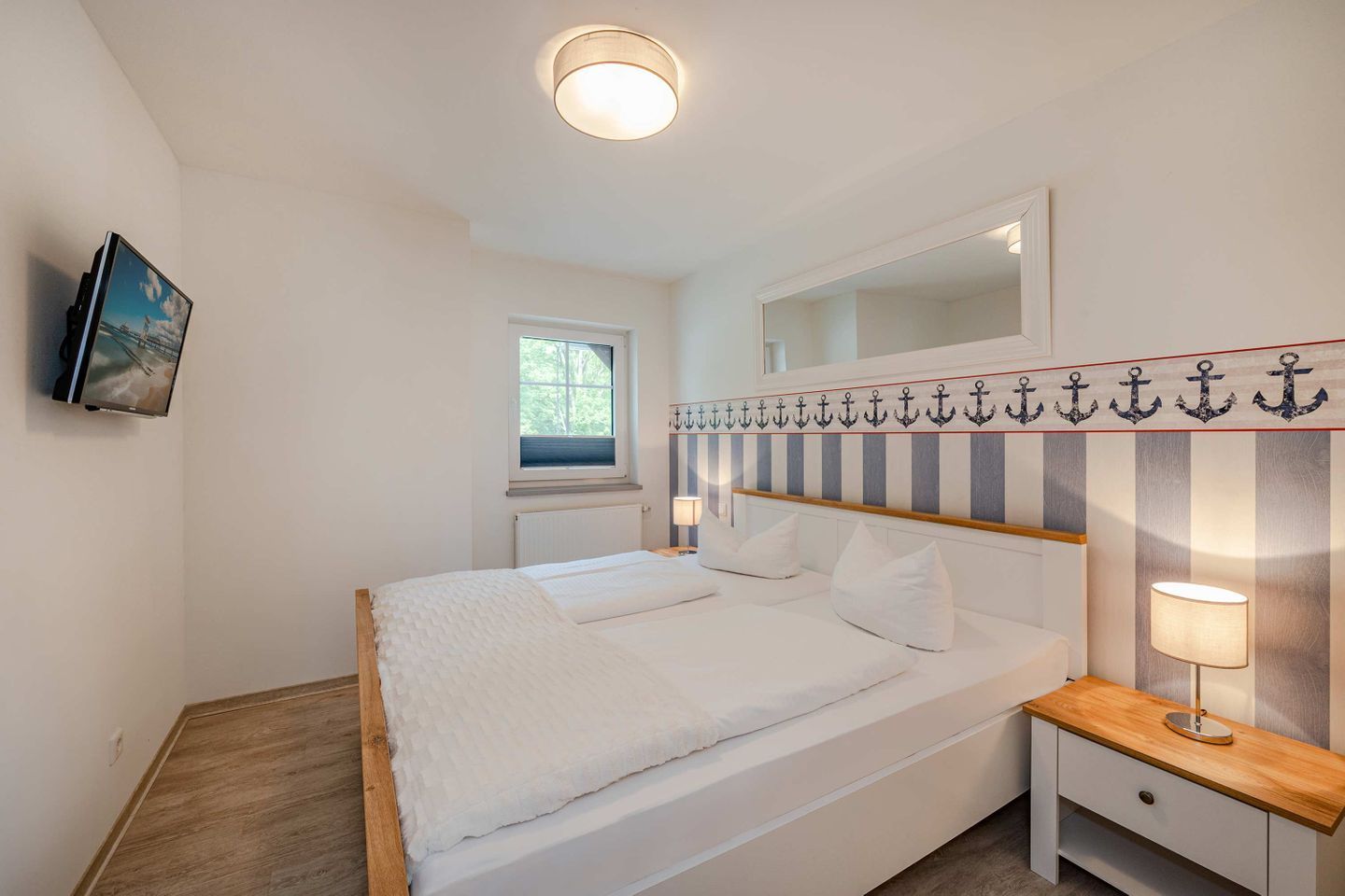 A bedroom with a bed, bedside table, lamp, window, and a wall-mounted TV.